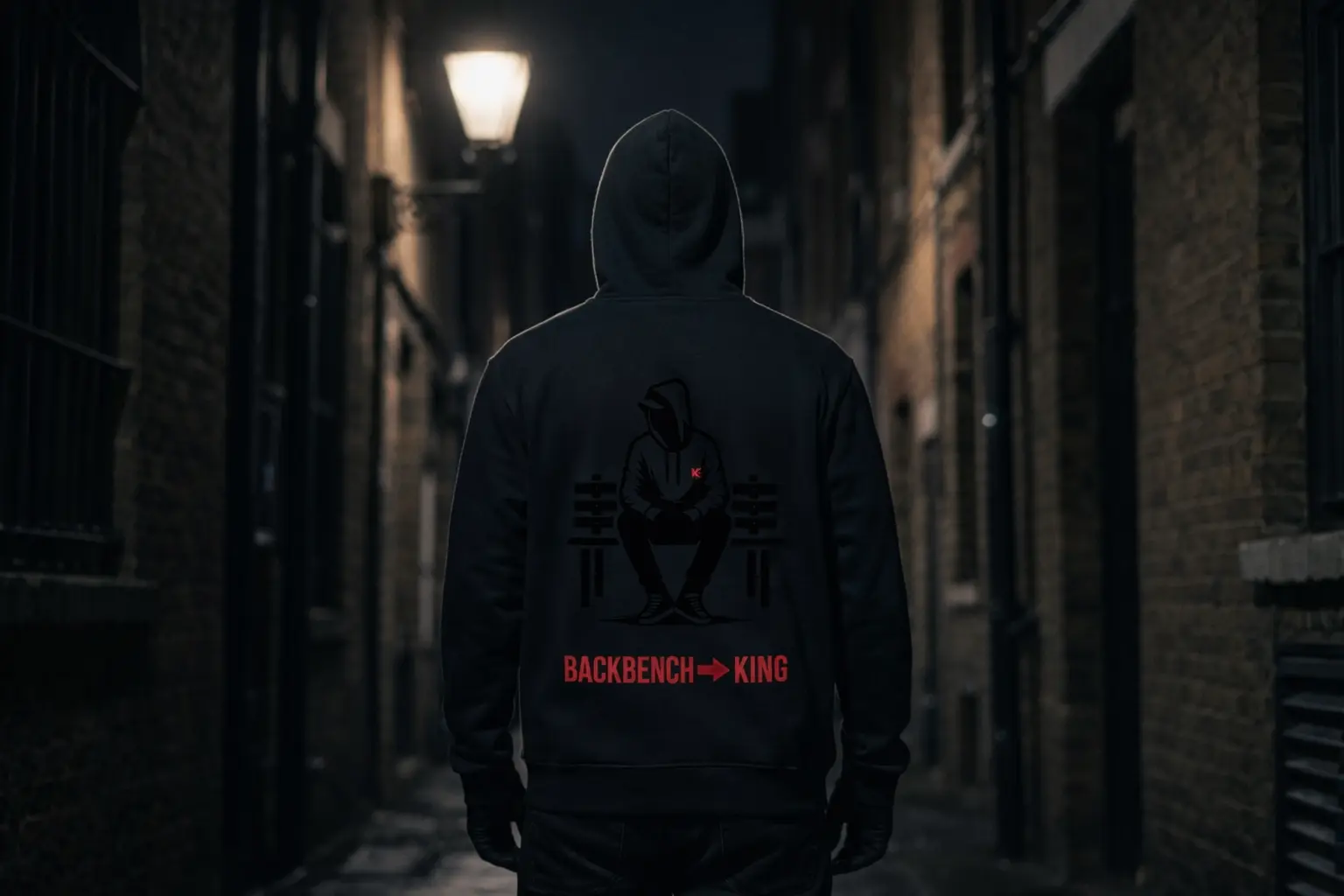 A figure in a black Backbencher hoodie walks down a wet London alley at night, Bench Guy print on the back